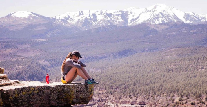 Girl, hugging her knees, sitting at the edge of a cliff with mountains in the background.
