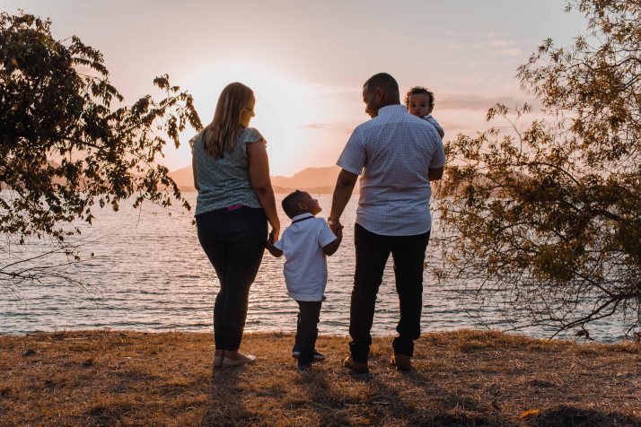 two parents standing holding the hand of a child between them and Dad holding the other child facing a body of water while the sun is setting